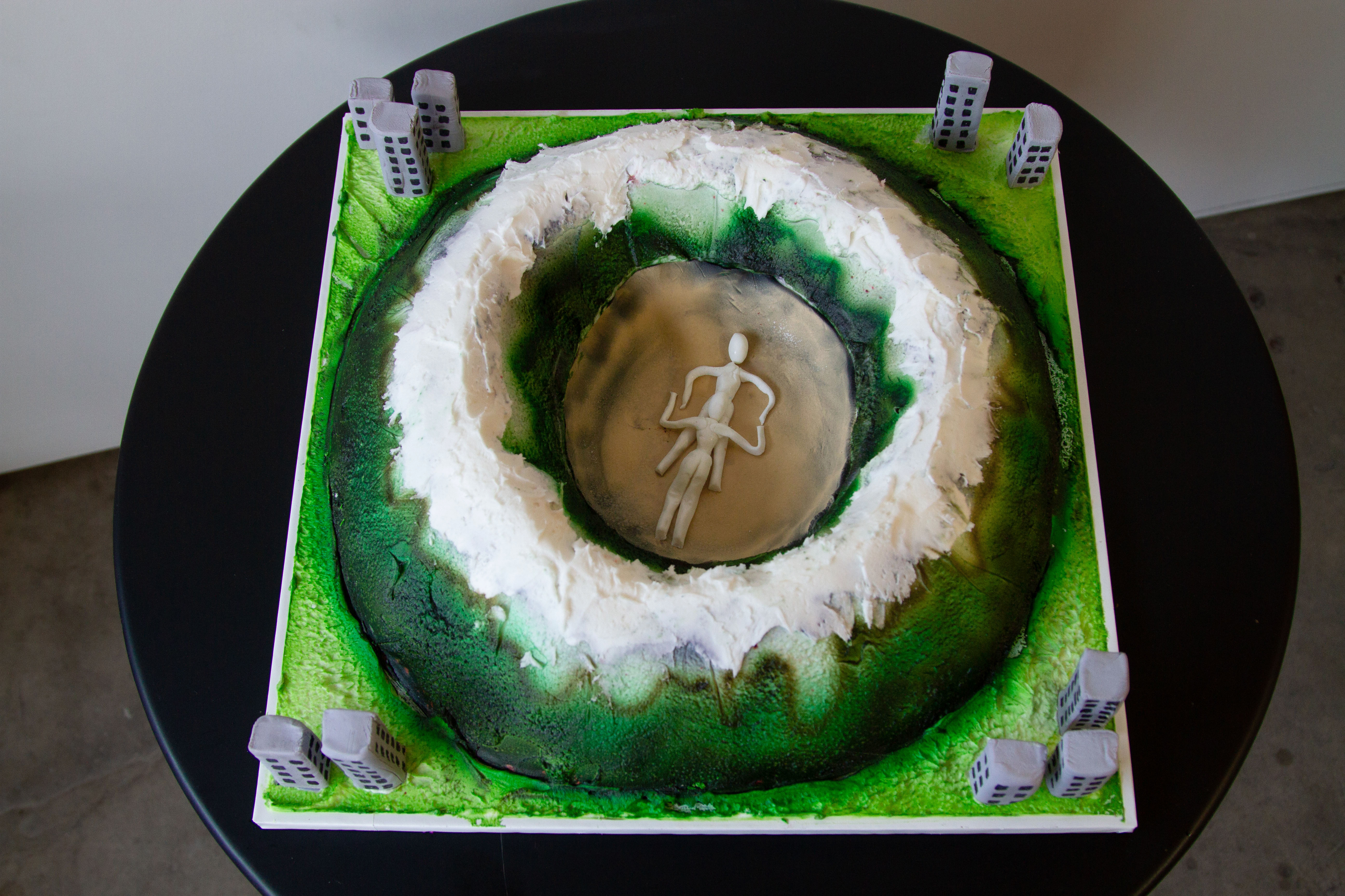 Ariel 3/4 view of a cake shaped like a mountainous crater with two figures engaged in felatio in the center surrounded by buildings on a black table